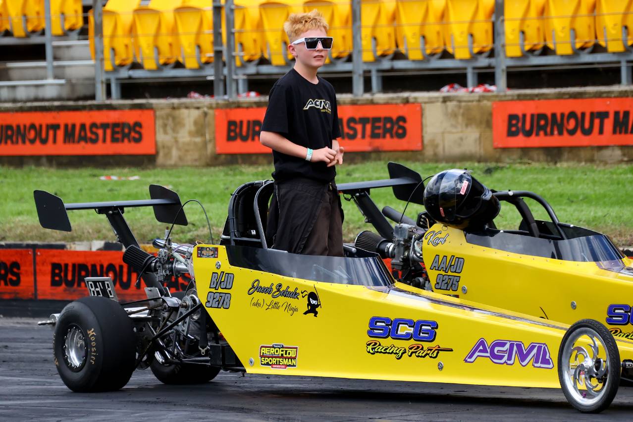 Jacob Schulz wearing sunglasses and standing in the cockpit of his yellow Junior Dragster, beside another yellow dragster.